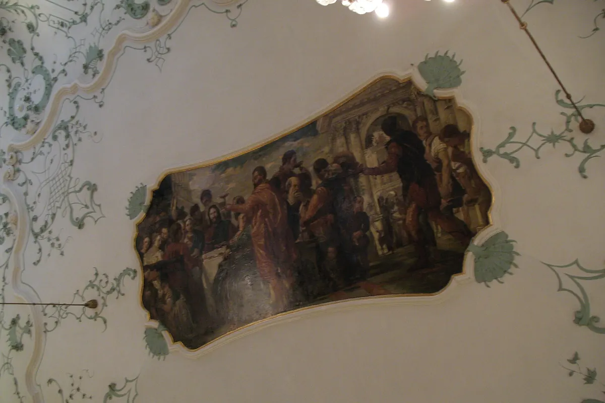 A painted panel of figures around a table set into a white Rococo ceiling with green scrollwork.