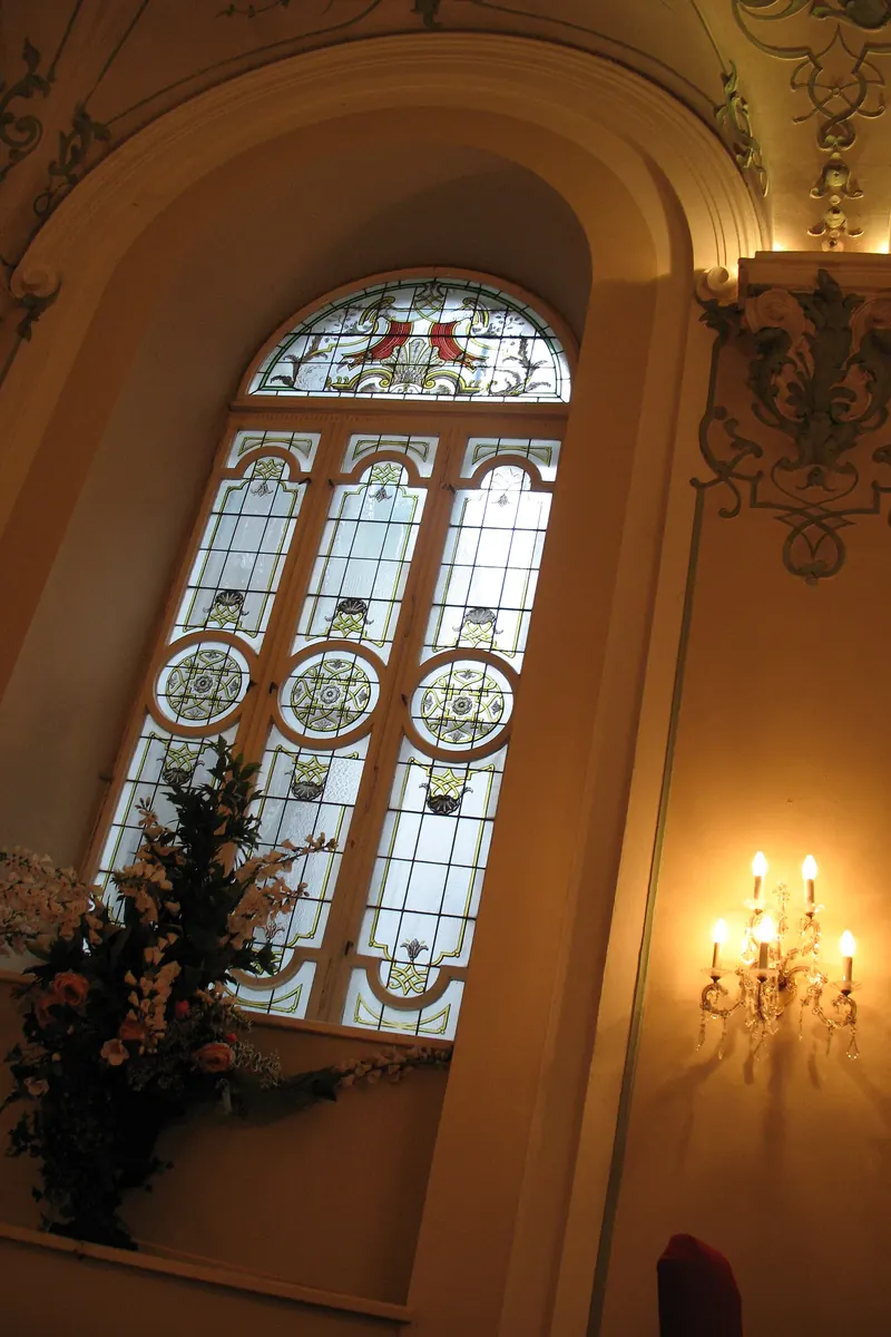 A tall arched stained-glass window in a cream wall with gilded stucco and a wall sconce, beside a flower arrangement.